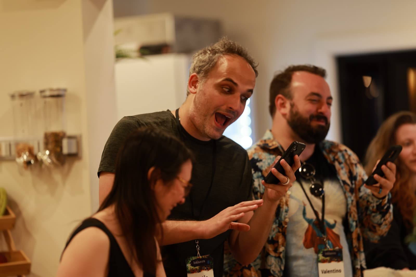 Three attendees laughing in a kitchen while looking at phones together.