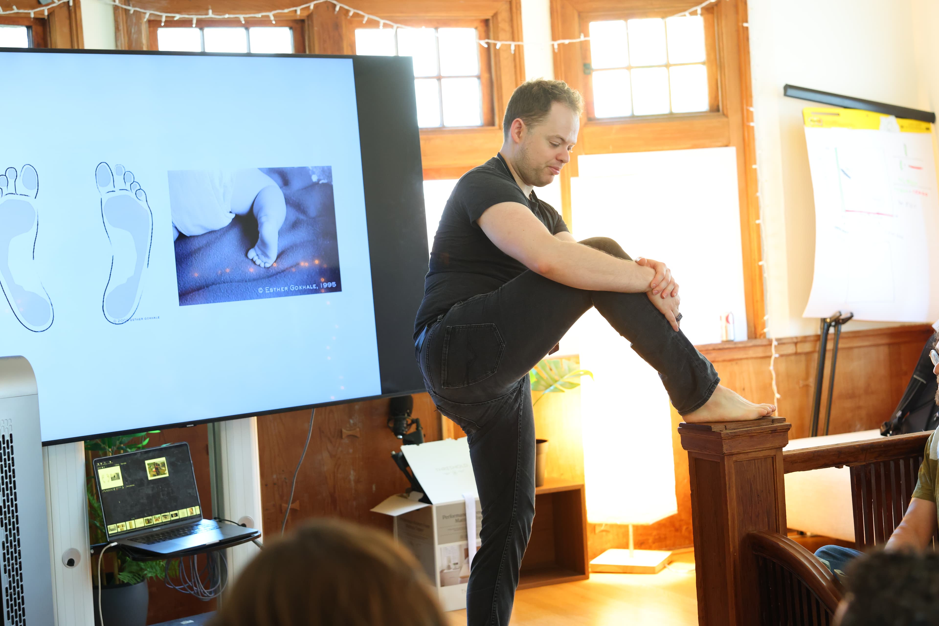 A speaker stands barefoot on a chair pedestal demonstrating posture.