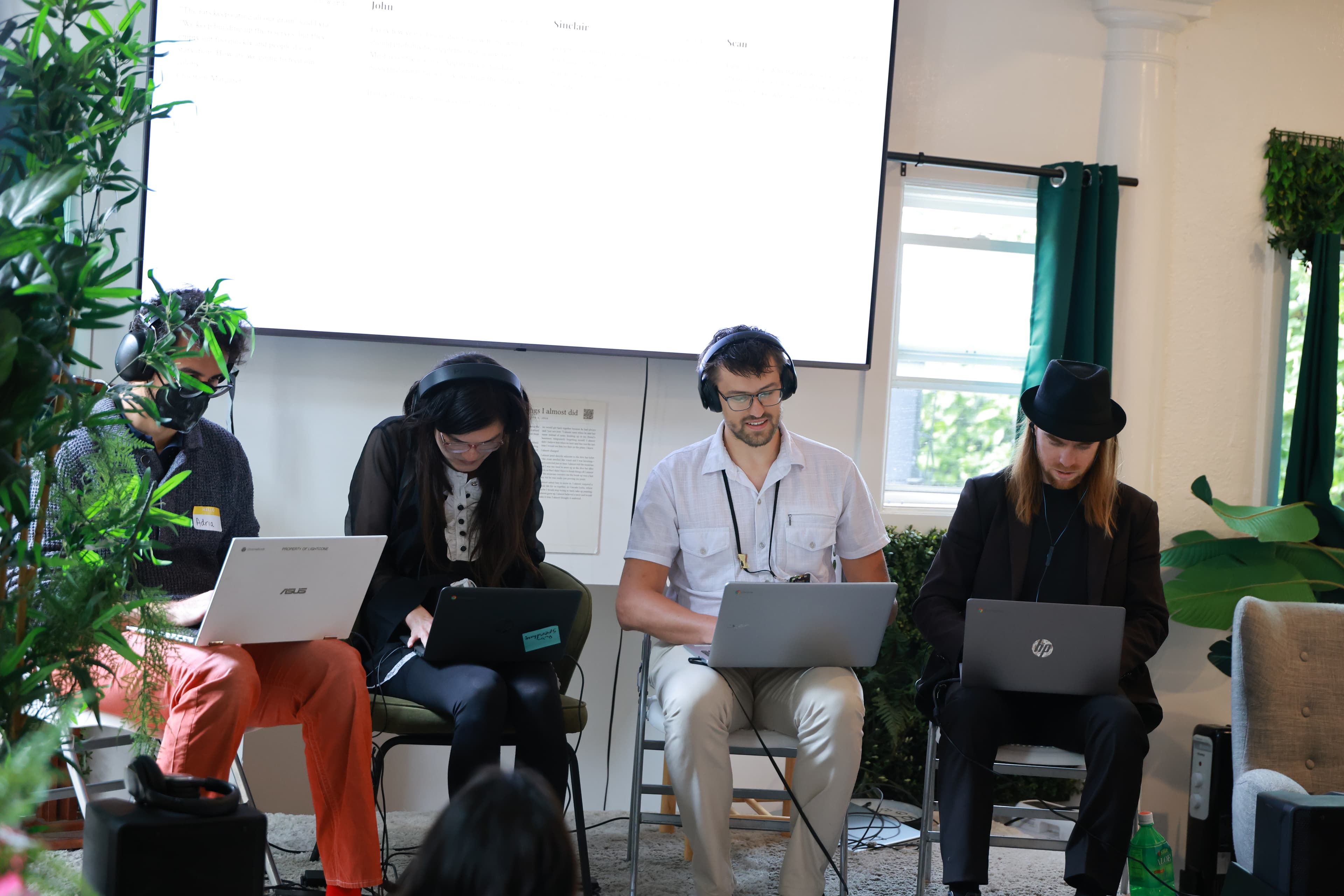 Four hosts seated at long tables on stage with laptops in front of an audience.