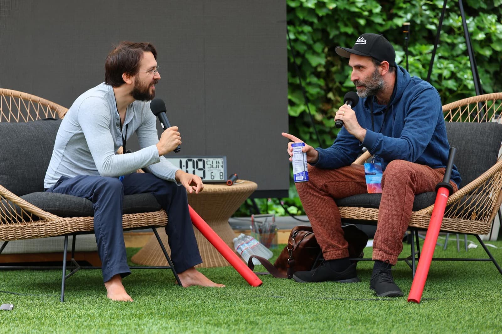 Two people seated across from each other in rattan chairs on the lawn, each holding a microphone.